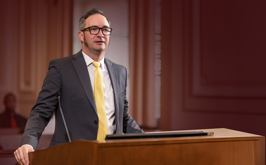 A man in a suit and yellow tie stands at a wooden podium in a courtroom, blurred people behind him, maroon gradient on the right.