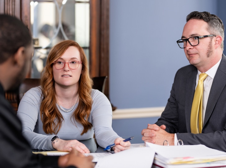 A red-haired woman with glasses speaks, holding a pen at a table with documents, sitting with two men in an office setting.
