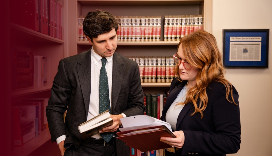 A man in a suit and a woman in business attire review documents together in a law library with bookshelves in the background.
