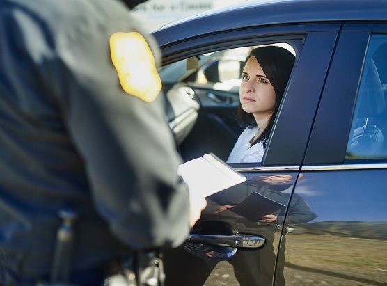 A woman in a car's driver seat looks up at a police officer outside her window, who is writing on a notepad.