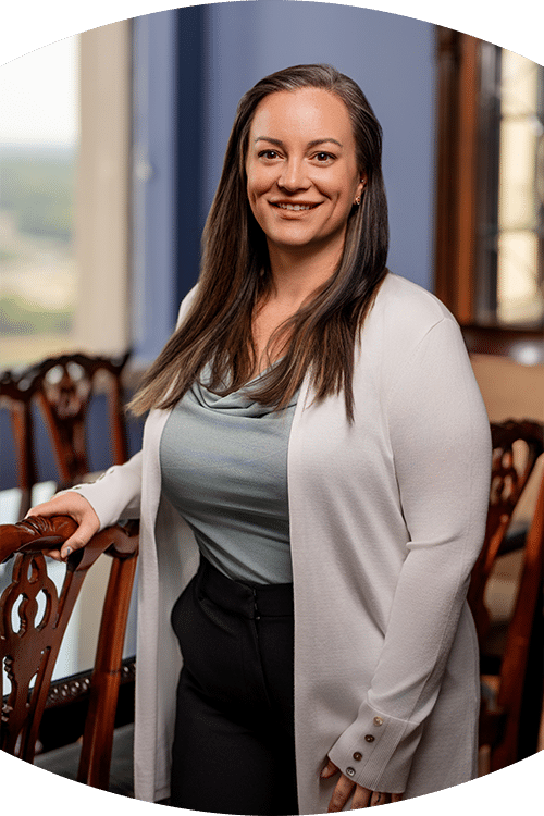 A woman with long brown hair in a blue top, white cardigan, and black pants stands smiling by a wooden chair in a bright room.
