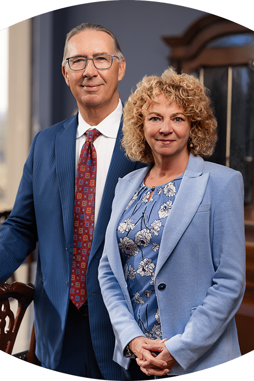 A man in a blue suit and red patterned tie stands next to a woman in a light blue blazer and floral blouse, both smiling indoors.