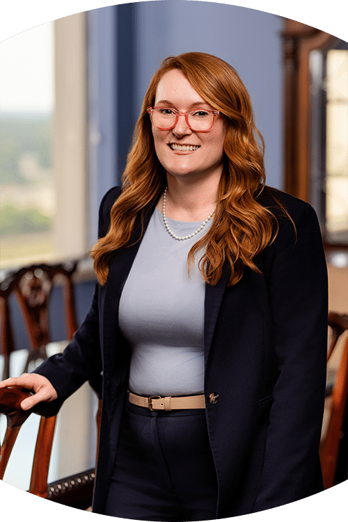 A woman with long red hair, red glasses, navy blazer, and pearls smiles in a formal room with wooden chairs and large windows.