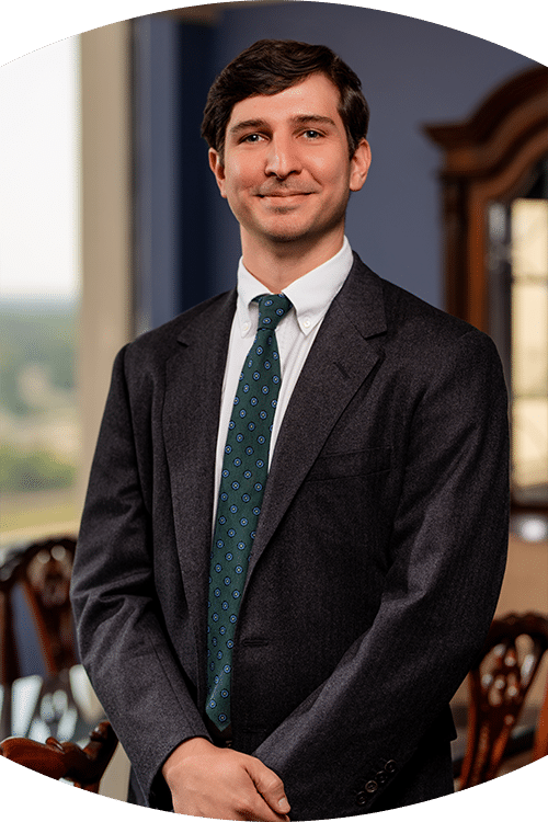 A man in a dark suit, white shirt, and green patterned tie stands indoors, smiling slightly with a window and wooden furniture behind him.