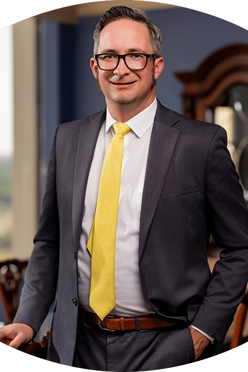 A man in glasses, gray suit, white shirt, and bright yellow tie stands indoors smiling. A wooden cabinet and window are behind him.