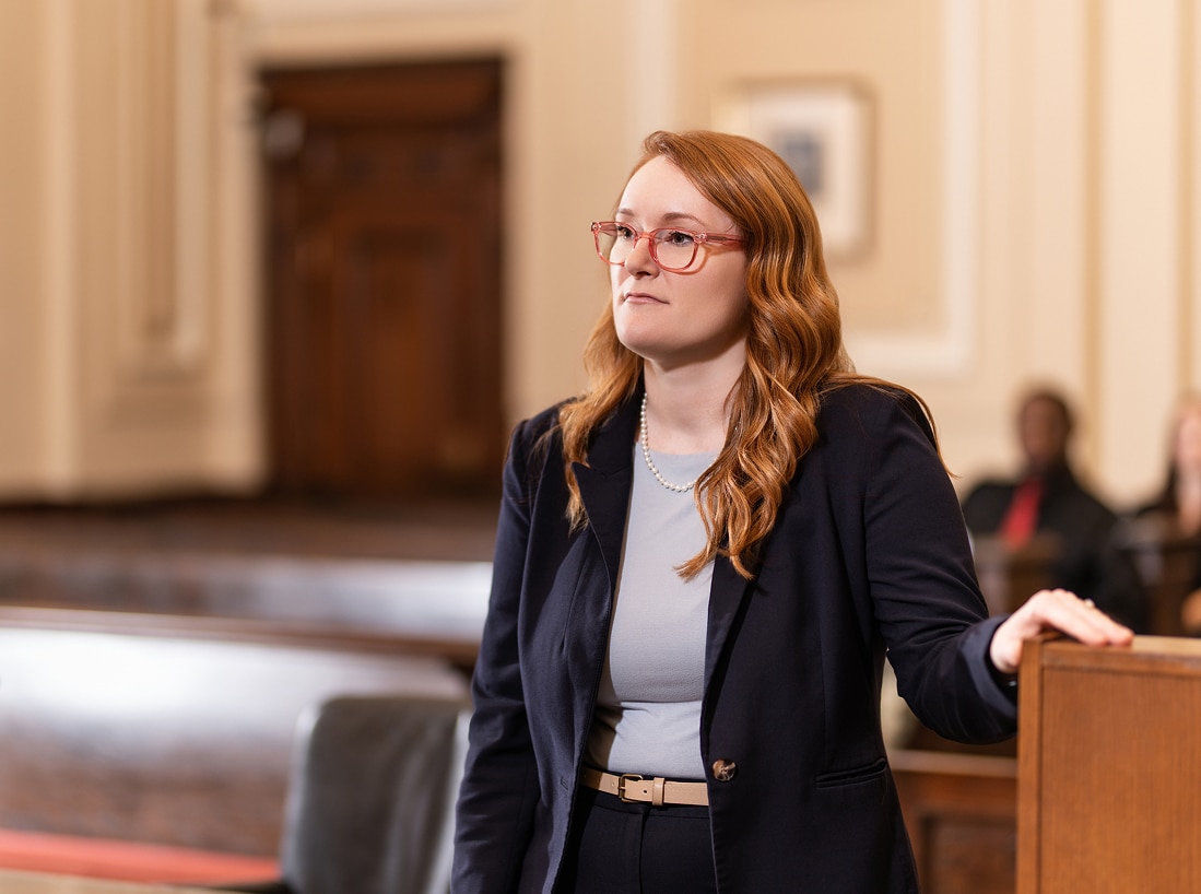 A woman with long red hair and glasses in a navy suit stands confidently at a courtroom podium; benches and figures blur behind her.