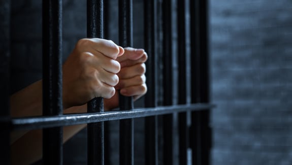 Hands firmly holding onto metal prison bars in a shadowy cell, dark brick wall visible behind, light barely illuminating the scene.