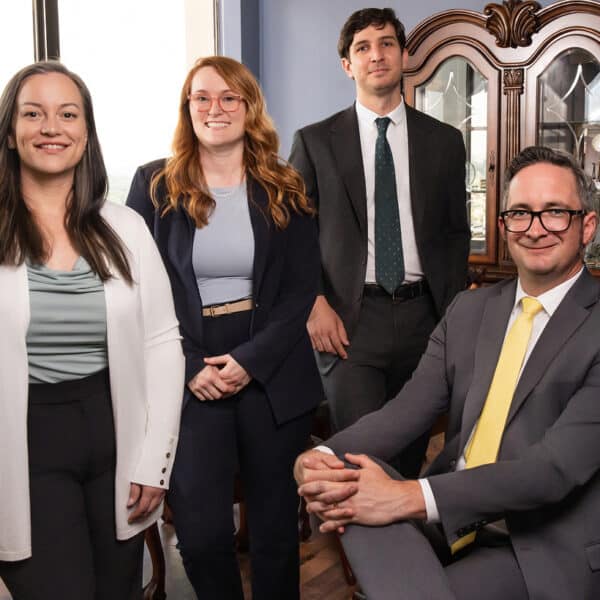 Four professionally dressed people in an office with a window and wood cabinet; three stand, one is seated and smiling at the camera.