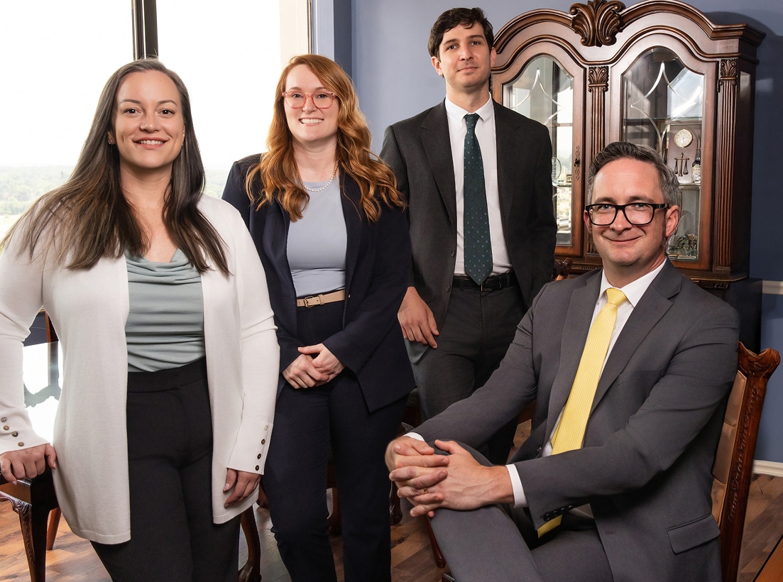 Four professionally dressed people in an office with a window and wood cabinet; three stand, one is seated and smiling at the camera.