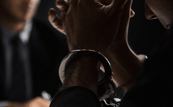 Close-up of a handcuffed person with clasped hands across from someone in a suit, likely during an interrogation in dim lighting.
