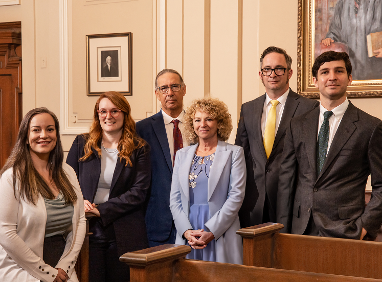 Six professionally dressed adults, three men and three women, pose in a wood-paneled room with art on cream-colored walls behind them.