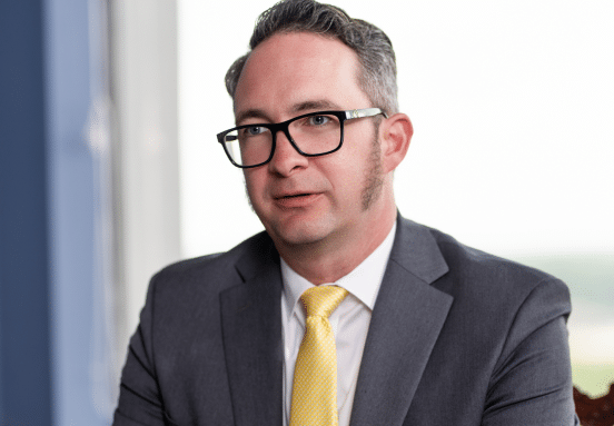 Man with short gray hair, glasses, and sideburns in a gray suit, white shirt, yellow tie sits indoors by a blurred window view.