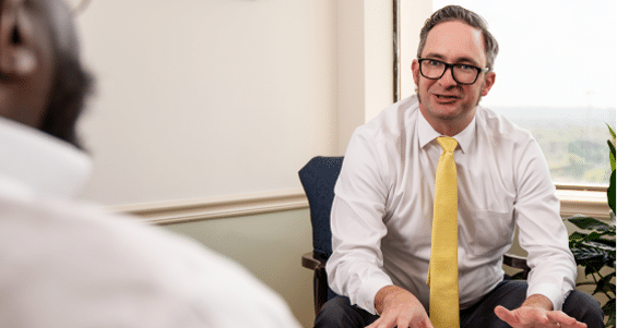 A man in glasses and a white shirt with a yellow tie sits in an office chair, talking to someone. A window and green plant are behind.