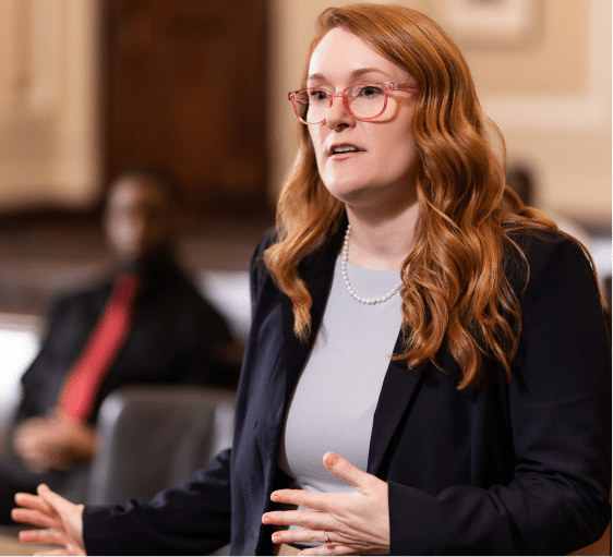 A woman with long red hair, glasses, navy blazer, and pearls gestures in a formal room. A man in a suit is blurred behind her.