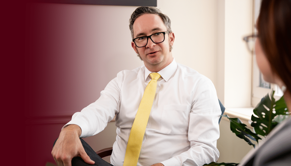A man in glasses, a white shirt, and yellow tie sits cross-legged, talking to someone in a bright office with plants and neutral walls.