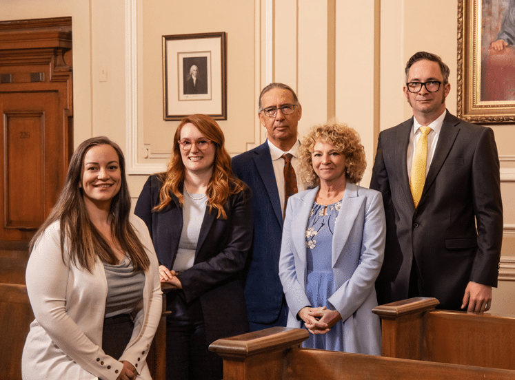 Five people in business attire pose for a group photo in a wood-paneled courtroom with framed portraits and beige walls behind them.