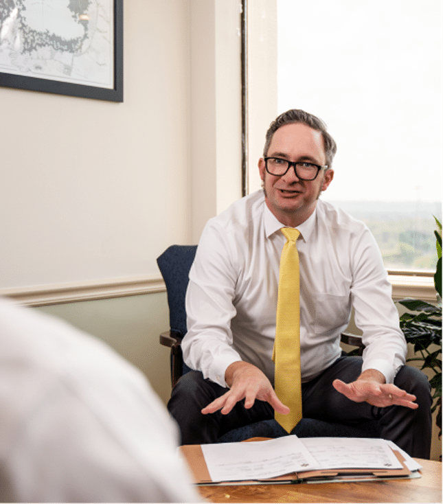 Man in a white shirt and yellow tie sits in an office chair, gesturing while talking. Papers are spread out on the table before him.
